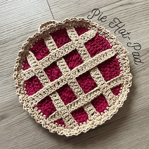 May include: A crocheted pie-shaped hot pad with a beige crust and a red filling. The hot pad is made with a woven pattern and has a scalloped edge.