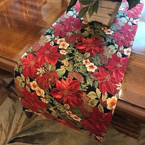 May include: A black table runner with a red and green floral pattern. The runner features poinsettia flowers, holly, and other winter greenery. The runner is on a wooden table with a vase of red berries and pine branches.