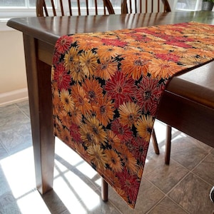 May include: A table runner with a floral pattern featuring orange, red, and yellow flowers on a black background. The runner is on a wooden table with two chairs.