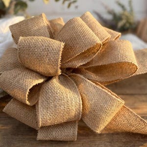 May include: A large, decorative bow crafted from tan, coarse-textured burlap fabric. The bow features multiple voluminous loops arranged in a circular, layered pattern, showcasing the distinct woven fibers and natural light brown color of the material.