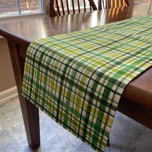 May include: A green and yellow plaid table runner with a white background. The runner is on a wooden table with two wooden chairs.