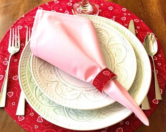 May include: A red placemat with white hearts and swirls, a white plate with a pink napkin, a silver fork and knife, and a glass of water.