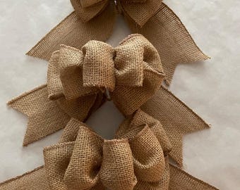 May include: Three decorative bows crafted from tan, woven burlap fabric. Each bow features a central knot with two loops on either side and two long, pointed tails extending downwards. The texture of the material is coarse and visible throughout.