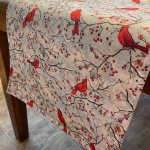 May include: A light blue table runner featuring a winter scene with red cardinals perched on branches with red berries. The runner is made of fabric and is approximately 60 inches long.