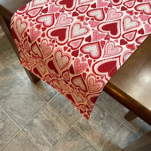 May include: A red and pink table runner with a heart pattern. The runner is made of fabric and has a white background.
