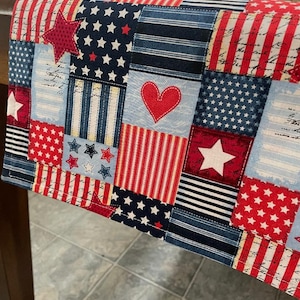 May include: Red, white, and blue patchwork table runner with stars and stripes. The runner is made of fabric and has a quilted look.