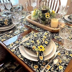 May include: A dining table setting featuring black placemats and napkins with a yellow sunflower pattern, white ceramic plates, clear crystal glassware, silver cutlery, and a wooden dough bowl centerpiece with candles and a decorative birdcage.