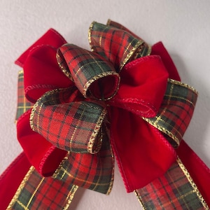 May include: A festive red bow with plaid ribbon accents. The bow features a classic plaid pattern in red, green, and gold, with a gold trim. The bow is made of a soft, velvet-like material, perfect for holiday decorations.