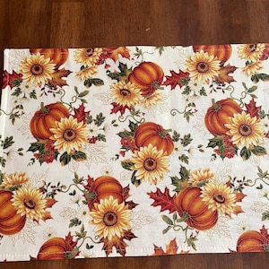 May include: A rectangular off-white table runner with a repeating pattern of orange pumpkins, yellow sunflowers, and red and green leaves. The runner is likely made of fabric and suitable for fall decor.