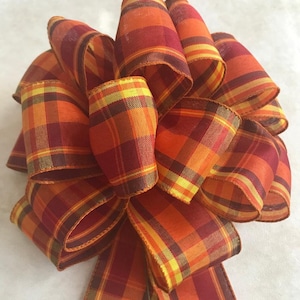 May include: A large decorative bow made from plaid ribbon in shades of orange, yellow, and burgundy. The bow has multiple loops and streamers, perfect for fall or autumn-themed decorations.
