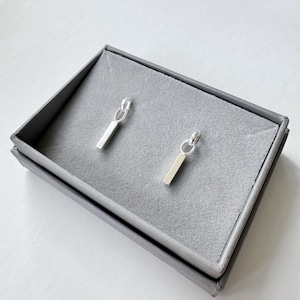 May include: A pair of silver earrings with a small bar pendant hanging from a loop. The earrings are displayed in a grey box.