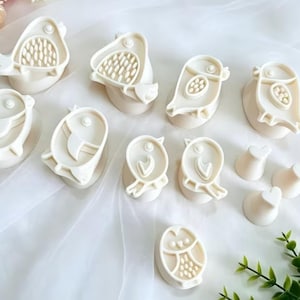 May include: A collection of off-white plastic cookie cutters and stamps featuring various bird shapes, including owls and songbirds, alongside small heart-shaped pieces, all arranged on a soft, light-colored fabric surface.