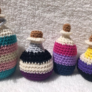 May include: Four crocheted bottles in different sizes, each with a different colour scheme representing a pride flag. The largest bottle is white, pink, and turquoise, the next is white, black, and purple, the next is pink, purple, and blue, and the smallest is yellow, white, purple, and black.