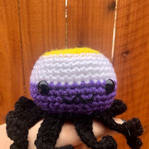 May include: A crocheted octopus plush toy with a purple, white and yellow body. The octopus has black crocheted tentacles.