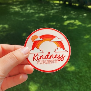 May include: A white round sticker with a red border. The sticker features a rainbow with clouds and a sun. The text "Kindness Counts" is written in red cursive lettering.