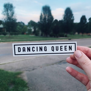 Puede incluir: Una pegatina blanca con texto negro que dice "Dancing Queen" sobre un fondo borroso de árboles y un camino pavimentado.