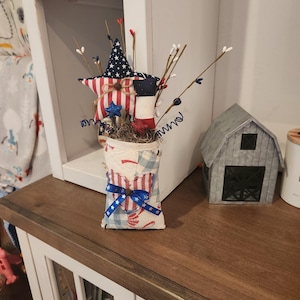 May include: A fabric-covered container featuring a red, white, and blue patchwork pattern with a blue ribbon bow. It holds a decorative arrangement of fabric stars, including one with white stars on blue and red stripes, alongside thin twigs and wire accents.