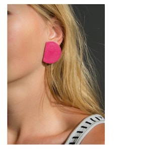 Könnte beinhalten: Nahaufnahme eines Ohrs mit einem großen, leuchtend pinkfarbenen, halbkreisförmigen Ohrring. Der Ohrring hat eine einfarbige, strukturierte Oberfläche. Die Person hat lange, blonde Haare und trägt ein schwarz-weiß gestreiftes Oberteil.
