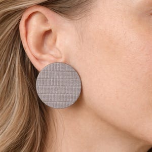 A close-up of a gray, circular stud earring with a textured, grid-like pattern. The earring is worn on the ear of a person with blonde hair. The earring is approximately 1.5 inches in diameter. Make a bold statement with these oversized grey raptile embossed leather stud earrings, designed for lovers of minimalist style with a modern urban edge. Crafted from exceptionally soft genuine leather, these large yet ultra-lightweight earrings offer striking presence without compromising comfort.