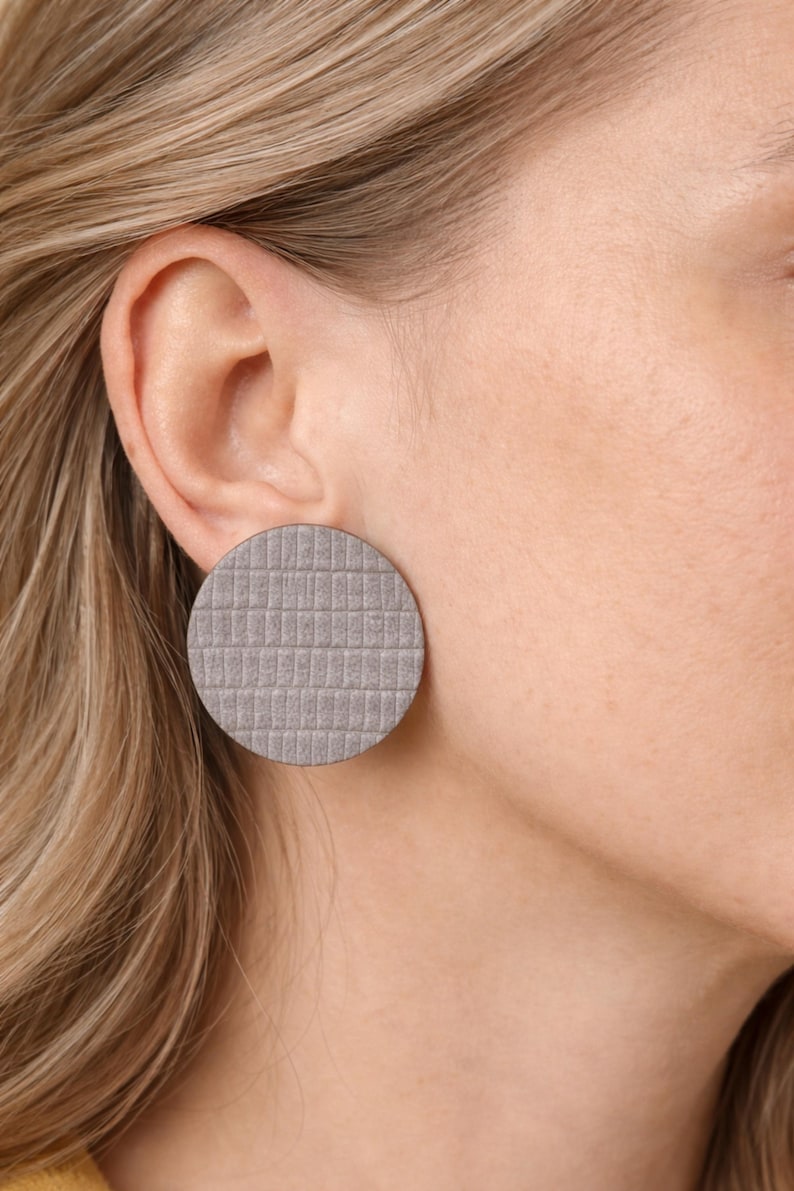 A close-up of a gray, circular stud earring with a textured, grid-like pattern. The earring is worn on the ear of a person with blonde hair. The earring is approximately 1.5 inches in diameter. Make a bold statement with these oversized grey raptile embossed leather stud earrings, designed for lovers of minimalist style with a modern urban edge. Crafted from exceptionally soft genuine leather, these large yet ultra-lightweight earrings offer striking presence without compromising comfort.