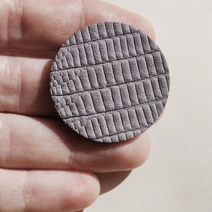 A round, textured, gray leather piece with a reptile scale pattern. The leather is a muted gray color, and the pattern is embossed, creating a tactile surface. The piece is held in a hand. Make a bold statement with these oversized grey raptile embossed leather stud earrings, designed for lovers of minimalist style with a modern urban edge. Crafted from exceptionally soft genuine leather, these large yet ultra-lightweight earrings offer striking presence without compromising comfort.