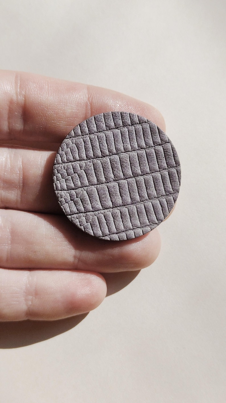 A round, textured, gray leather piece with a reptile scale pattern. The leather is a muted gray color, and the pattern is embossed, creating a tactile surface. The piece is held in a hand. Make a bold statement with these oversized grey raptile embossed leather stud earrings, designed for lovers of minimalist style with a modern urban edge. Crafted from exceptionally soft genuine leather, these large yet ultra-lightweight earrings offer striking presence without compromising comfort.