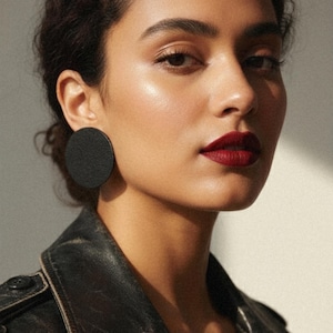 May include: A woman with dark hair and red lipstick wearing a black leather jacket and large, circular black earrings. The image is a close-up, focusing on the face and upper body. The earrings are a prominent feature.