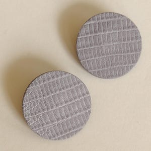 Two round, gray, textured earrings with a reptile-skin pattern. The earrings are a neutral color and could be worn with many outfits. Make a bold statement with these oversized grey raptile embossed leather stud earrings, designed for lovers of minimalist style with a modern urban edge. Crafted from exceptionally soft genuine leather, these large yet ultra-lightweight earrings offer striking presence without compromising comfort.