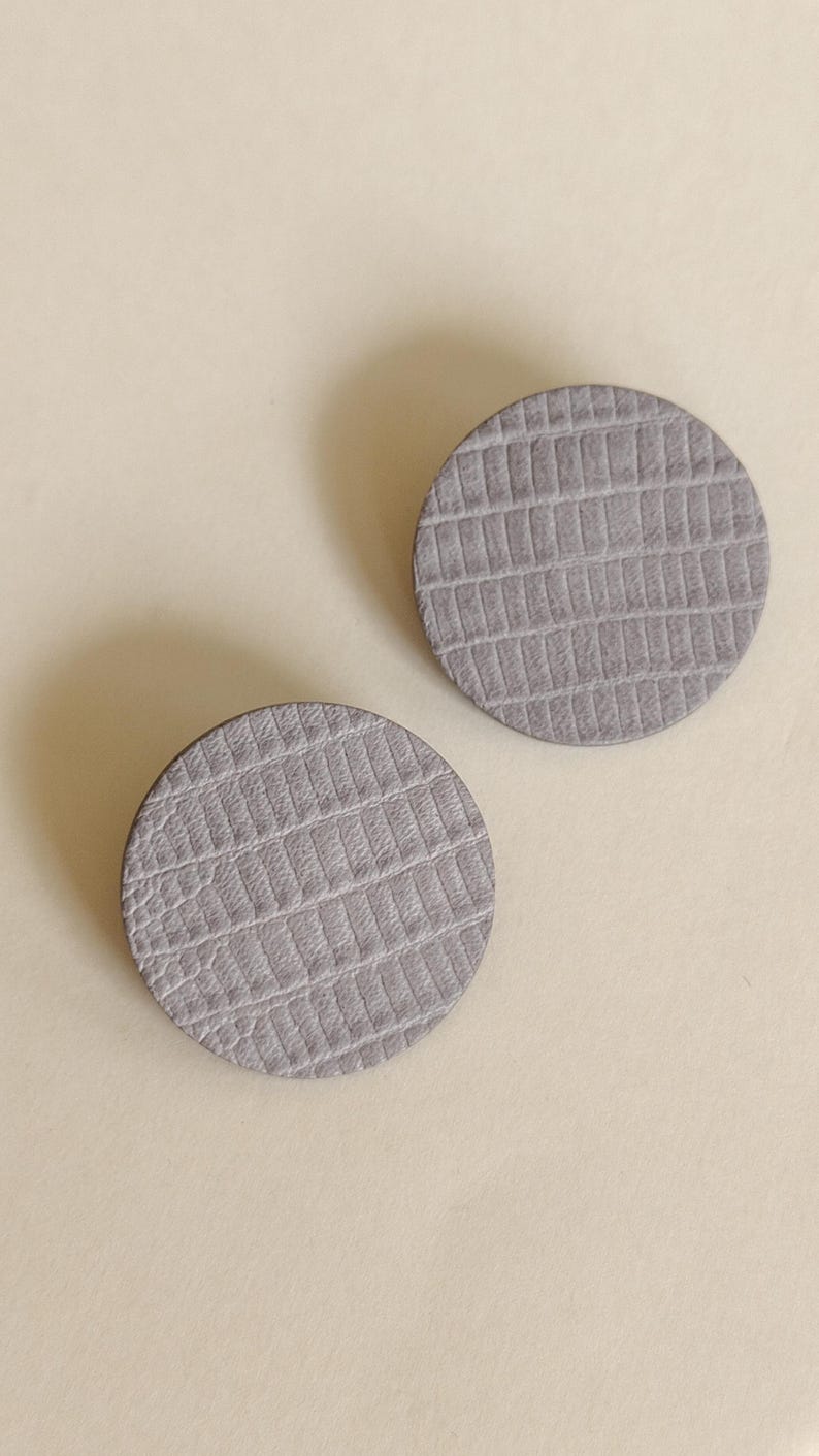 Two round, gray, textured earrings with a reptile-skin pattern. The earrings are a neutral color and could be worn with many outfits. Make a bold statement with these oversized grey raptile embossed leather stud earrings, designed for lovers of minimalist style with a modern urban edge. Crafted from exceptionally soft genuine leather, these large yet ultra-lightweight earrings offer striking presence without compromising comfort.