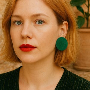 Könnte beinhalten: Eine Frau mit heller Haut und rotem Lippenstift trägt ein dunkelgrünes Oberteil und große, runde, waldgrüne Ohrringe. Ihr kurzes, rötlich-blondes Haar umrahmt ihr Gesicht. Der Hintergrund ist eine strukturierte, hellbraune Wand.