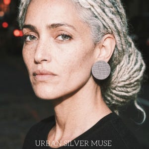 A gray hair woman wearing a pair of gray circular earrings. The earrings have a textured surface - animal print, snake pattern leather studs, geometric modern look, modern urban muse with gray dreadlocks. Make a bold statement with these oversized grey raptile embossed leather stud earrings, designed for lovers of minimalist style with a modern urban edge.  These large yet ultra-lightweight earrings offer striking presence without compromising comfort.