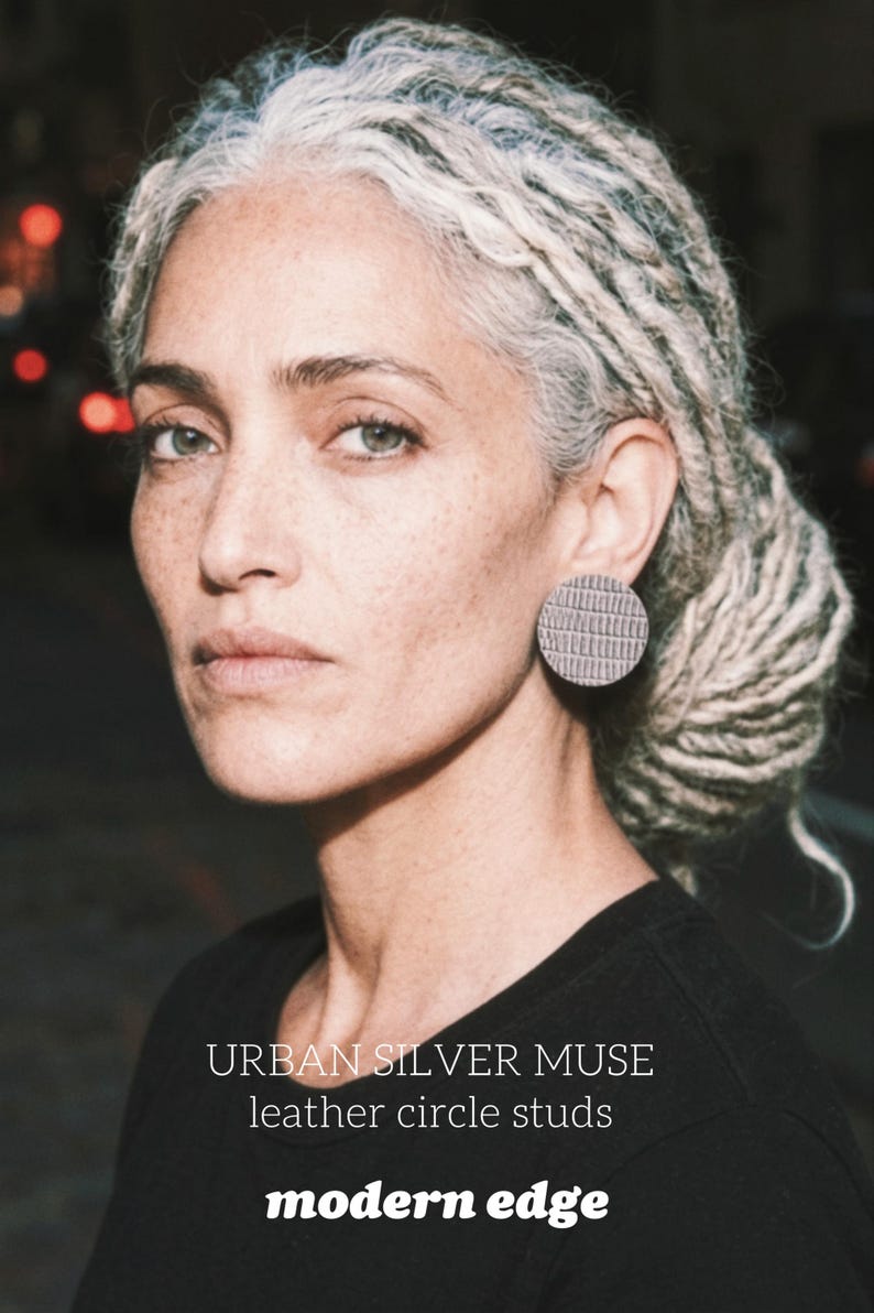 A gray hair woman wearing a pair of gray circular earrings. The earrings have a textured surface - animal print, snake pattern leather studs, geometric modern look, modern urban muse with gray dreadlocks. Make a bold statement with these oversized grey raptile embossed leather stud earrings, designed for lovers of minimalist style with a modern urban edge.  These large yet ultra-lightweight earrings offer striking presence without compromising comfort.