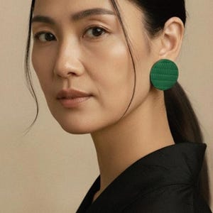 Op de afbeelding: Een vrouw draagt een zwarte top en een paar groene, cirkelvormige oorbellen. De oorbellen hebben een textuur. De vrouw heeft donker haar dat in een paardenstaart is vastgebonden.