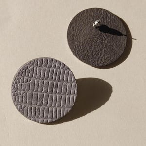 K&ouml;nnte beinhalten: Zwei graue Lederohrringe. Einer hat ein strukturiertes, krokodilartiges Muster. Der andere ist einfarbig grau mit einem Stecker zum Durchstechen. Die Ohrringe sind kreisf&ouml;rmig.