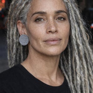May include: A woman with long, gray dreadlocks and a black t-shirt. She is wearing a large, round, gray earring. The woman has freckles and is looking directly at the camera.