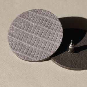 Close-up of two round, textured leather earrings. One is light gray with a reptile-like pattern. Make a bold statement with these oversized grey raptile embossed leather stud earrings, designed for lovers of minimalist style with a modern urban edge. Crafted from exceptionally soft genuine leather, these large yet ultra-lightweight earrings offer striking presence without compromising comfort.