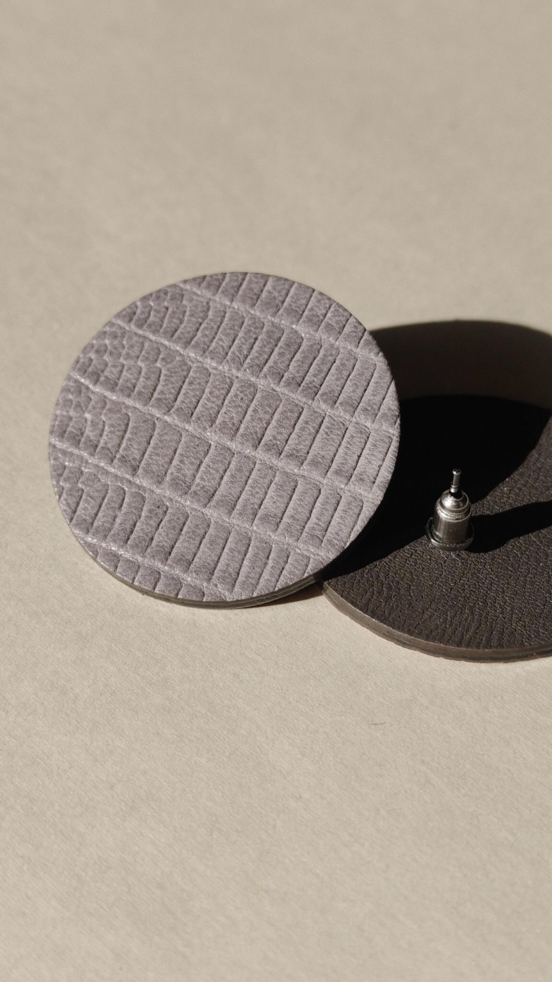 Close-up of two round, textured leather earrings. One is light gray with a reptile-like pattern. Make a bold statement with these oversized grey raptile embossed leather stud earrings, designed for lovers of minimalist style with a modern urban edge. Crafted from exceptionally soft genuine leather, these large yet ultra-lightweight earrings offer striking presence without compromising comfort.