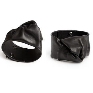 Puede incluir: Dos pulseras de cuero negro. Cada pulsera presenta un diseño anudado en la parte delantera. Los puños se aseguran con un cierre de botón a presión. Las pulseras se muestran sobre un fondo blanco.