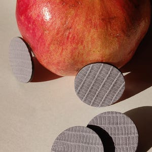 A ripe, red pomegranate with a textured surface, next to several gray, circular leather earrings. Make a bold statement with these oversized grey raptile embossed leather stud earrings, designed for lovers of minimalist style with a modern urban edge. Crafted from exceptionally soft genuine leather, these large yet ultra-lightweight earrings offer striking presence without compromising comfort.
