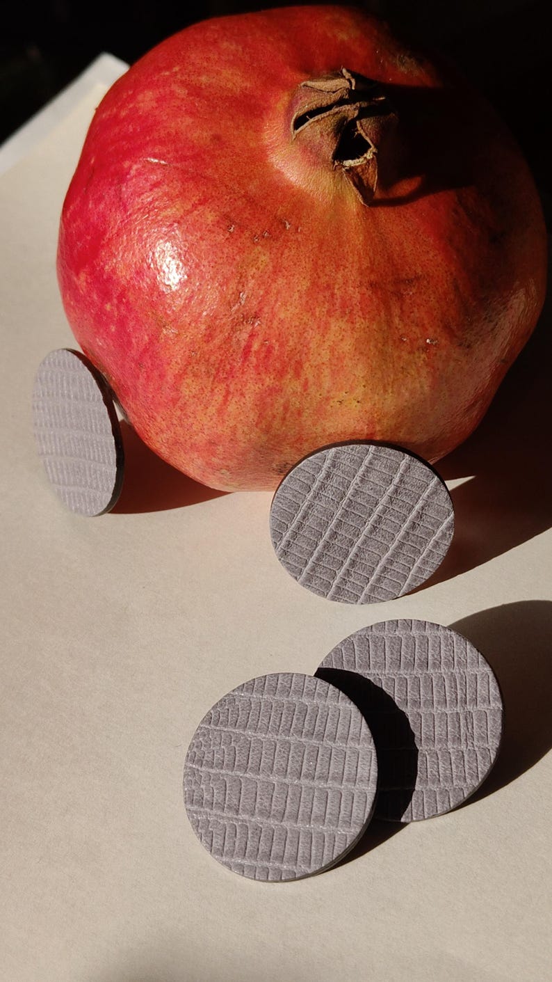 A ripe, red pomegranate with a textured surface, next to several gray, circular leather earrings. Make a bold statement with these oversized grey raptile embossed leather stud earrings, designed for lovers of minimalist style with a modern urban edge. Crafted from exceptionally soft genuine leather, these large yet ultra-lightweight earrings offer striking presence without compromising comfort.