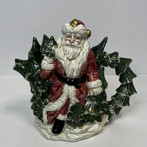May include: A ceramic teapot shaped like Santa Claus holding a Christmas tree. The teapot is decorated with green and gold details and has a white base.