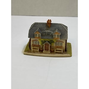 Puede incluir: Una réplica en miniatura de cerámica de un pub, "The Oak Inn", con un tejado de tejas grises, pequeñas ventanas y una chimenea. El edificio tiene una fachada color crema, una base verde y una puerta marrón. El texto "THE OAK INN" es visible.