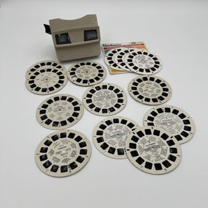 May include: A vintage View-Master viewer with a collection of circular image reels. The viewer is a light tan color, and the reels are white with black text and images. The image reels are arranged around the viewer.