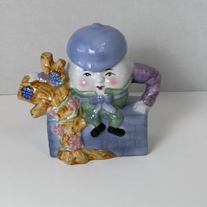 May include: A ceramic figurine of Humpty Dumpty, featuring a white egg-shaped body, a blue cap, and a green and purple outfit. The figurine is perched on a gray brick wall, with a brown tree branch adorned with flowers.