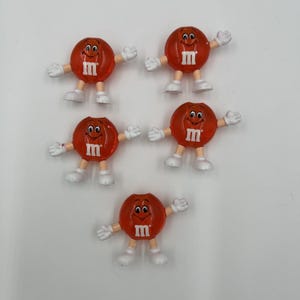 May include: Five red candy character figurines with white hands and feet. Each figure has a smiling face and the letter "m" on its front. The figures are arranged on a white surface.