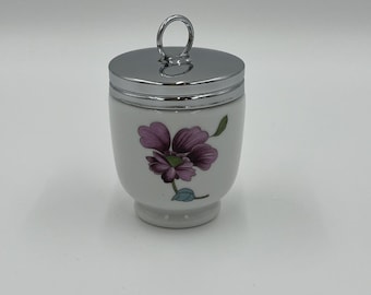 May include: White ceramic egg cup with a silver-colored lid and handle. The egg cup features a purple floral design. The lid is shiny and reflective. The egg cup is designed to hold a single egg.