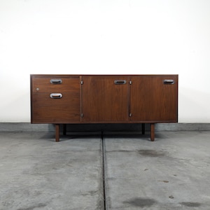 May include: A mid-century modern wooden sideboard with a dark brown finish. It features two drawers with silver handles on the left and two cabinet doors with silver handles on the right. The sideboard is supported by four short, tapered legs.