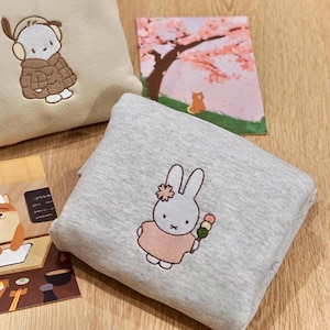 May include: A light grey sweatshirt with an embroidered cartoon bunny holding dango. A beige sweatshirt with an embroidered cartoon dog wearing a coat and earmuffs is also visible. Two illustrated cards are also in the image.