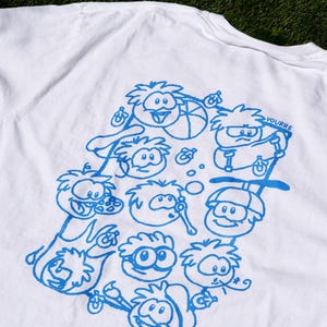 May include: A white t-shirt featuring a blue graphic print on the back. The design consists of multiple cartoon characters in various activities, including playing with a ball, skateboarding, and blowing bubbles, with the text VOURRE printed near the top right.
