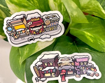 Puede incluir: Dos pegatinas con ilustraciones de dibujos animados de edificios con tejados cubiertos de nieve. Una pegatina muestra una tienda de mascotas, un escenario y una pizzería. La otra pegatina muestra una cafetería, un club nocturno y una tienda de regalos.